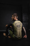BJJ athlete wearing the olive green Bone Breaking Jiu-Jitsu rashguard, seated on the training mat beside an MMA cage.